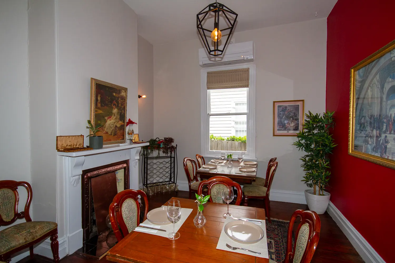 Elegant indoor dining room with vintage wooden chairs, framed artwork, a red feature wall and a set table by a window.