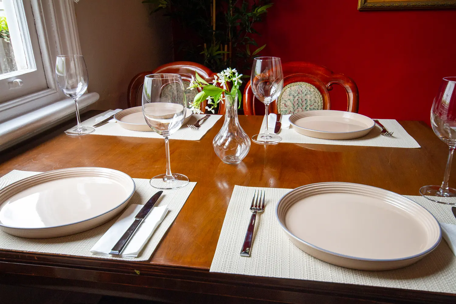 Close-up of a wooden dining table set with plates, cutlery, wine glasses and a small vase of flowers.
